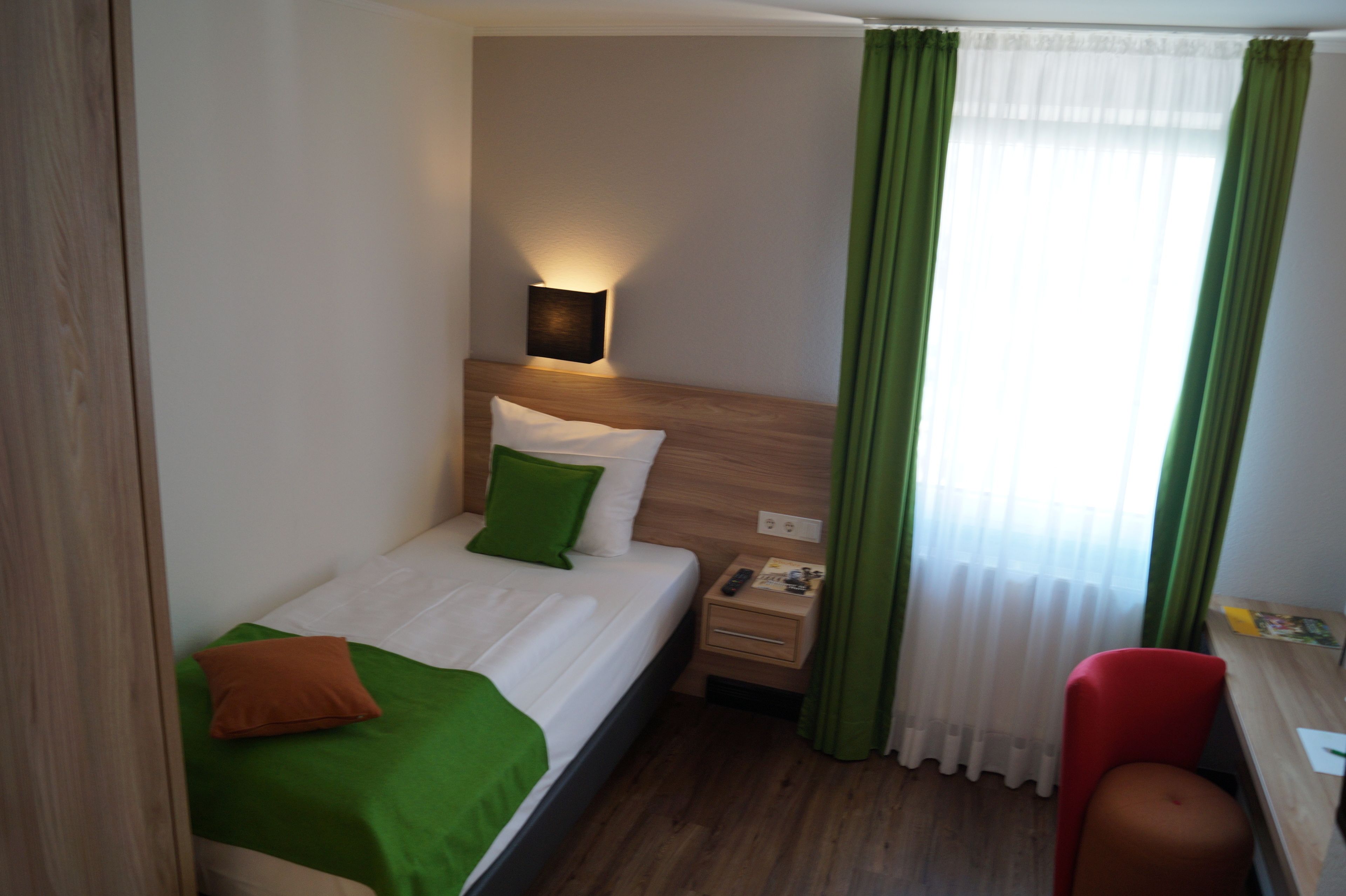 A hotel room with a bed, green and white bedding, a bedside table with a lamp, a nightstand, a chair, and a window with green curtains.