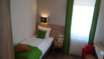 A hotel room with a bed, green and white bedding, a bedside table with a lamp, a nightstand, a chair, and a window with green curtains.