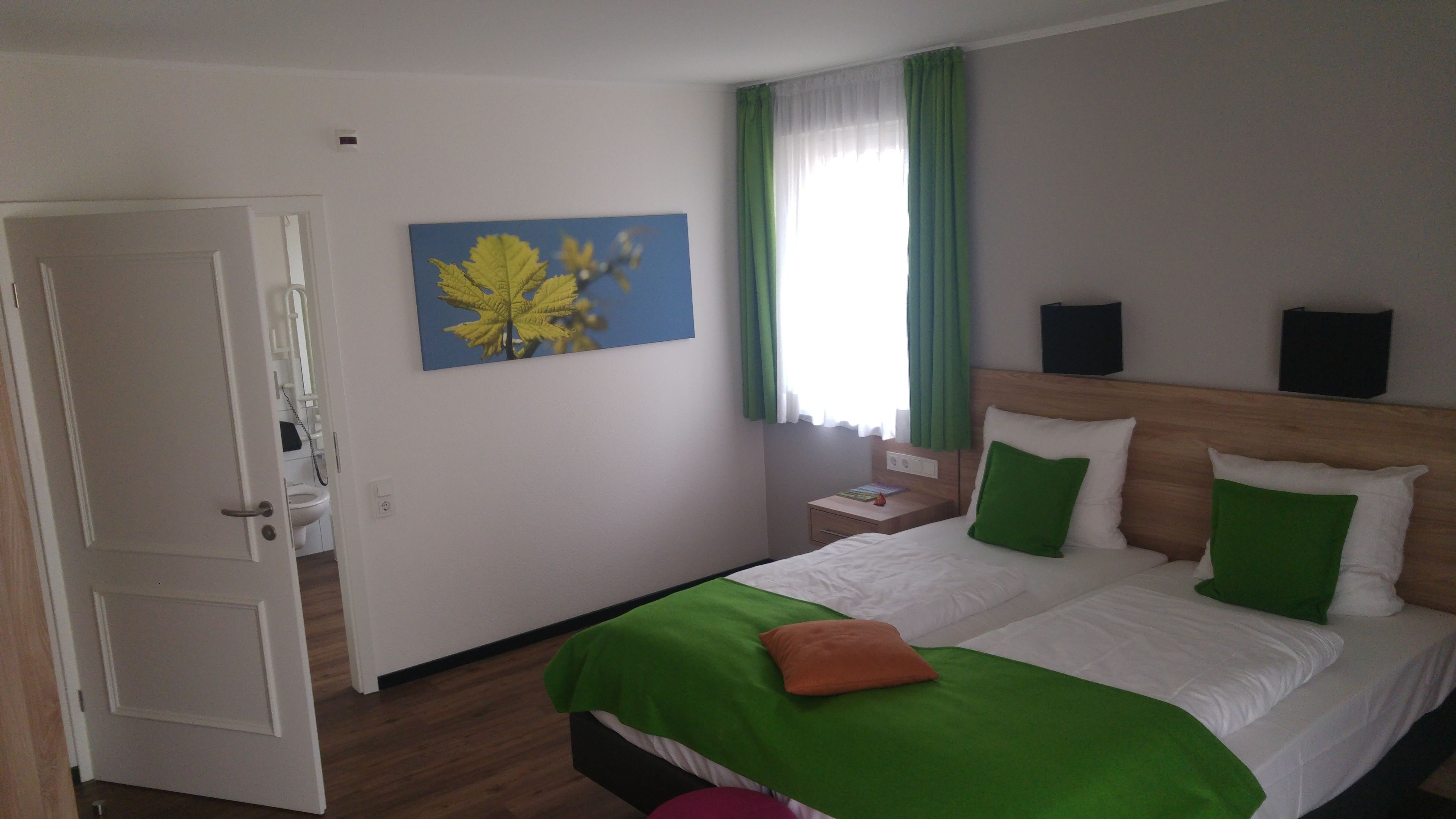 Habitación de hotel con una cama, ropa de cama verde y blanca, un cuadro con hojas, una mesita de noche y una ventana con cortinas.