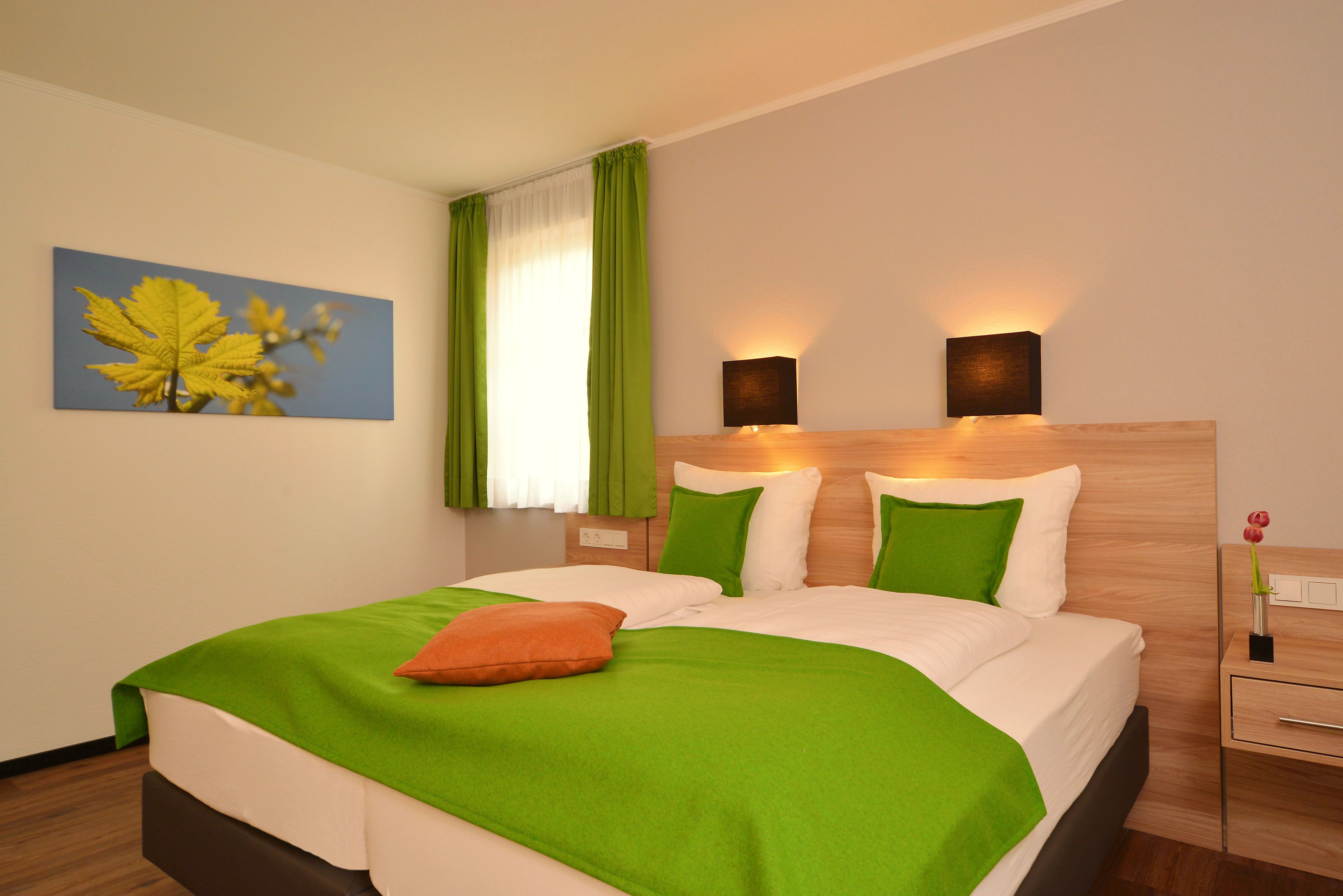 Une chambre d’hôtel moderne avec un grand lit, une literie verte et blanche, un tableau représentant des feuilles accroché au mur et deux lampes fixées au mur.