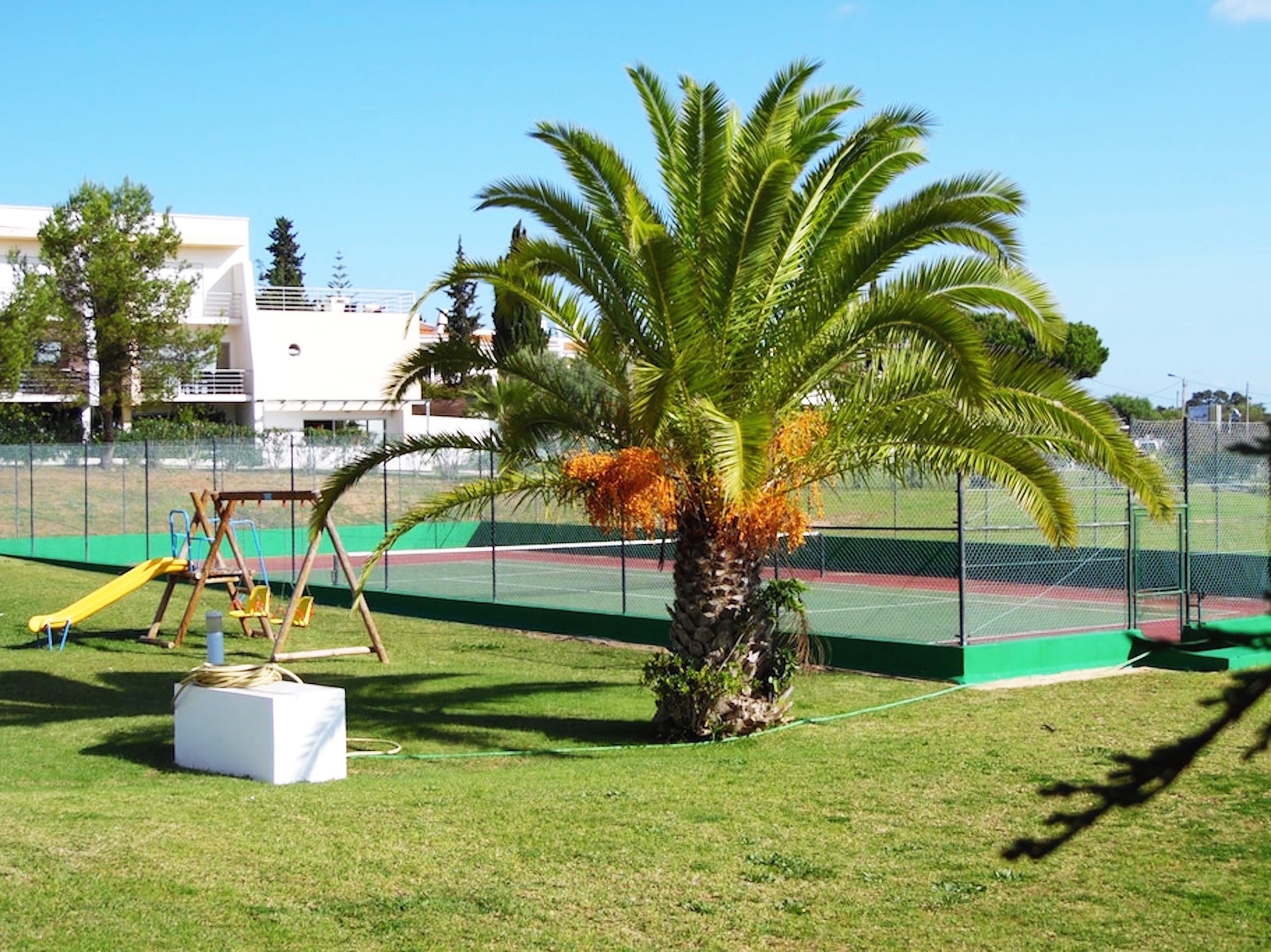 A playground with a swing set and a slide, surrounded by a fenced area with a palm tree.