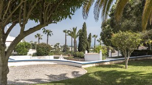 A cobblestone courtyard with a curved white wall, palm trees, and a view of the sea.