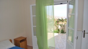 Une chambre à coucher avec un lit, une table de chevet en bois, un rideau vert transparent et une vue sur un jardin par une porte ouverte.
