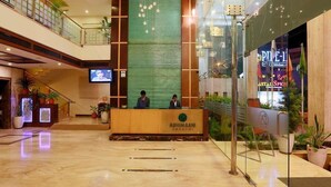 Et hotellobby med en receptionsdisk, to siddende personer, et fjernsyn og et skilt med teksten "Pine-1".