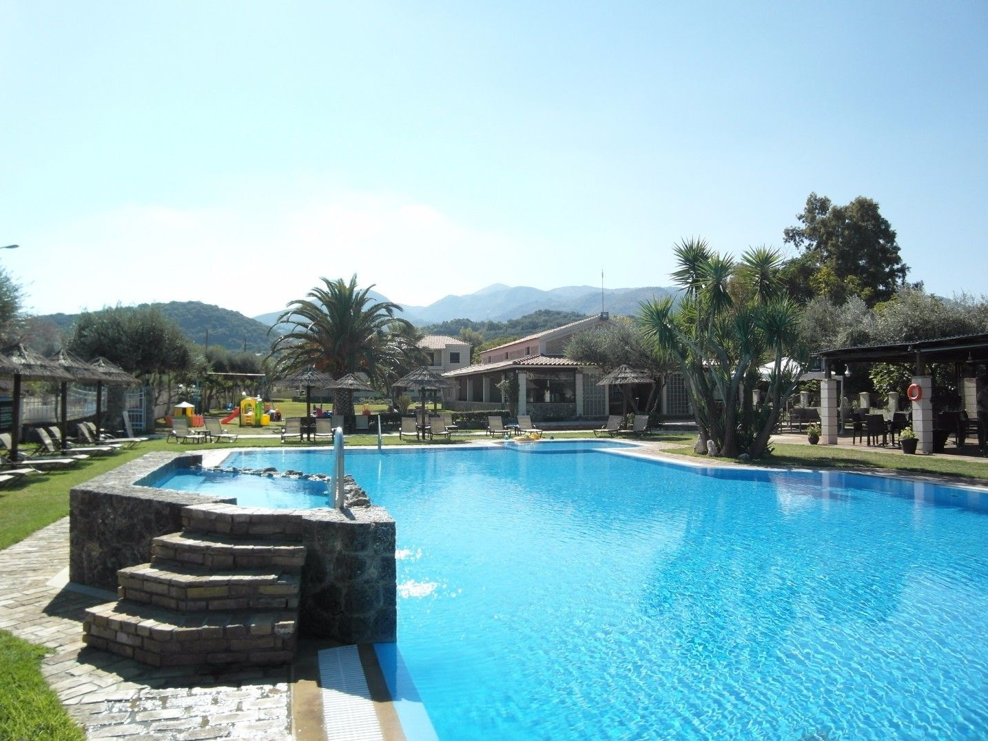 Una piscina con una escalera de piedra que da acceso a ella, rodeada de sillas de descanso y sombrillas, con montañas al fondo.