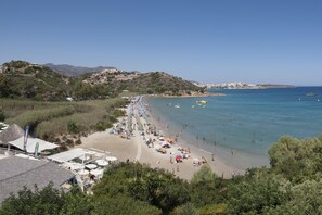A coastal beach with a long stretch of beach umbrellas and sun loungers, a clear blue sea, and buildings in the background. - Almiros Apartments (Corfu)