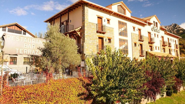 Hotel con ristorante e terrazza bar, facciata in pietra, numerosi balconi e montagne sullo sfondo.
