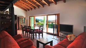 Un salon spacieux avec un canapé rouge, un coin repas comprenant une table et des chaises, un téléviseur et une vue sur un paysage verdoyant grâce à de grandes portes coulissantes.