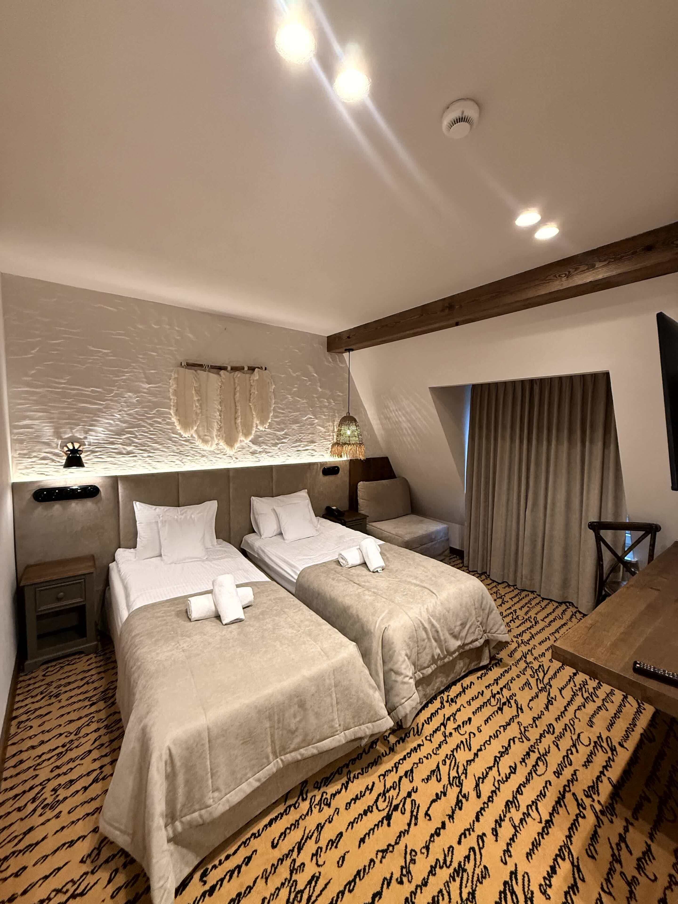 A hotel room with two beds, a desk, a chair, a large window with curtains, and a textured wall.