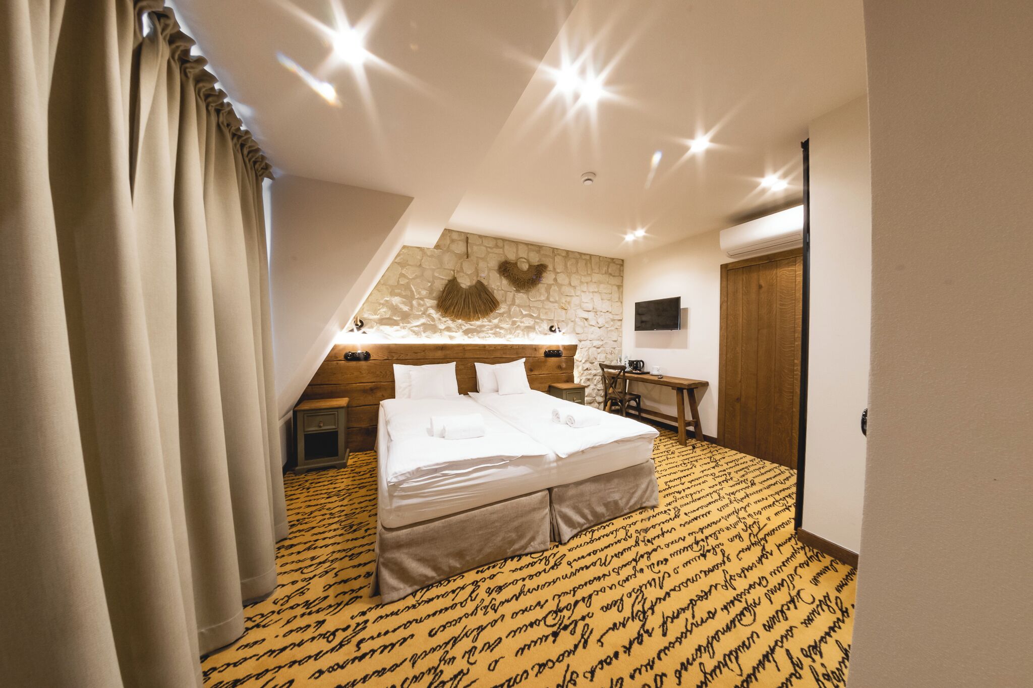 A hotel room with a large bed, a stone wall, a wooden desk, and a small window.