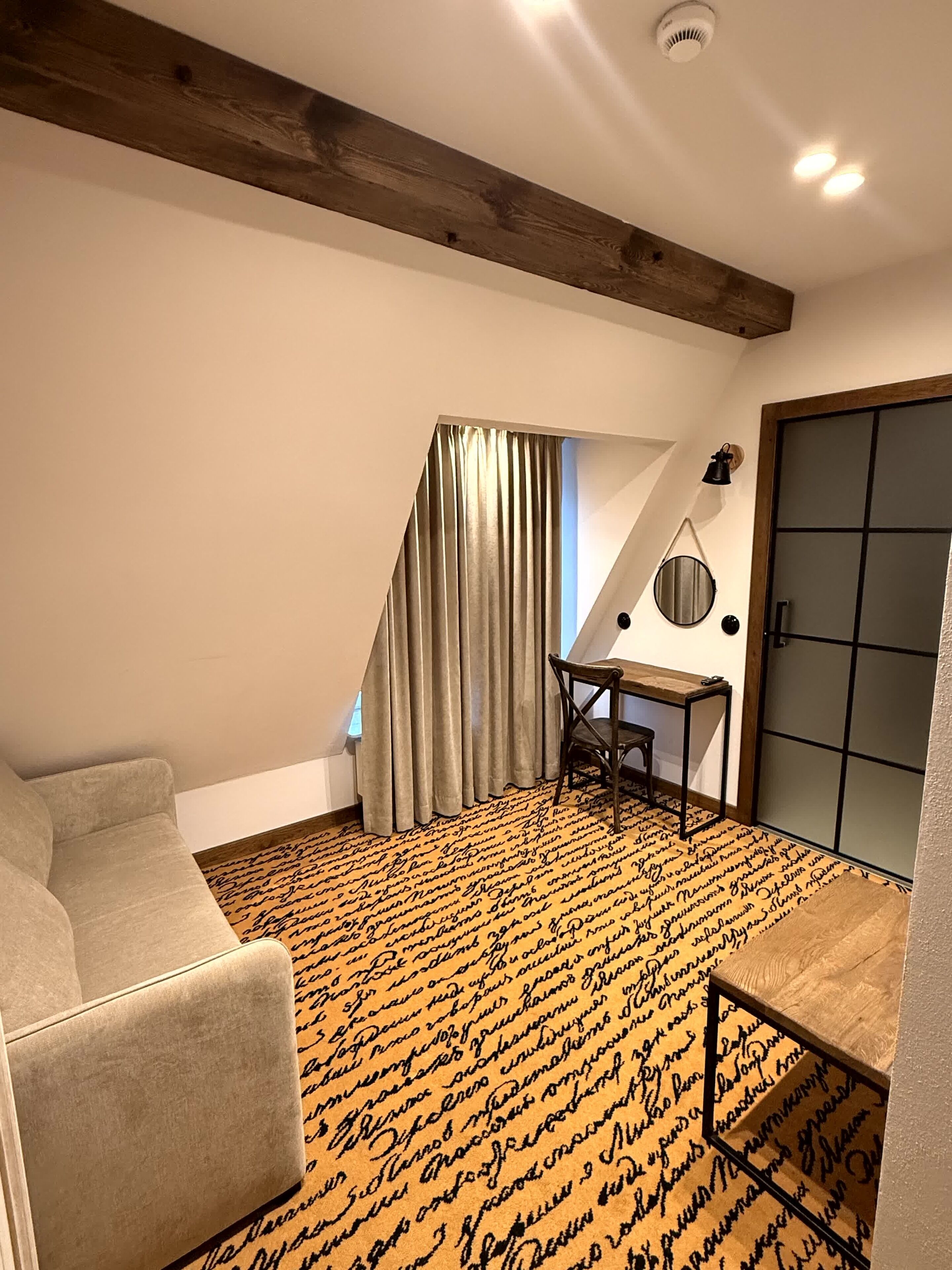 A room with a sloped ceiling, a desk with a chair, a window with curtains, and a patterned carpet.