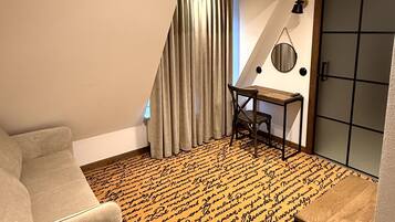 A room with a sloped ceiling, a desk with a chair, a window with curtains, and a patterned carpet.