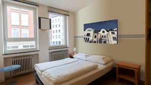 Un dormitorio con una cama doble, dos ventanas, un radiador, una mesita y un cuadro en la pared.