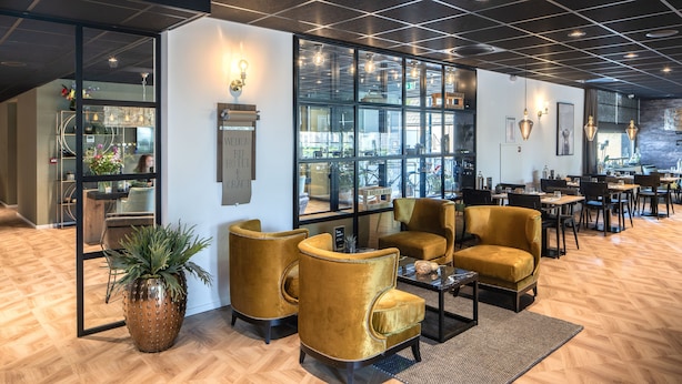 A modern hotel lobby with a wooden herringbone floor, large windows, and a seating area with gold velvet armchairs and a coffee table. - Hotel 46 (Wintelre)