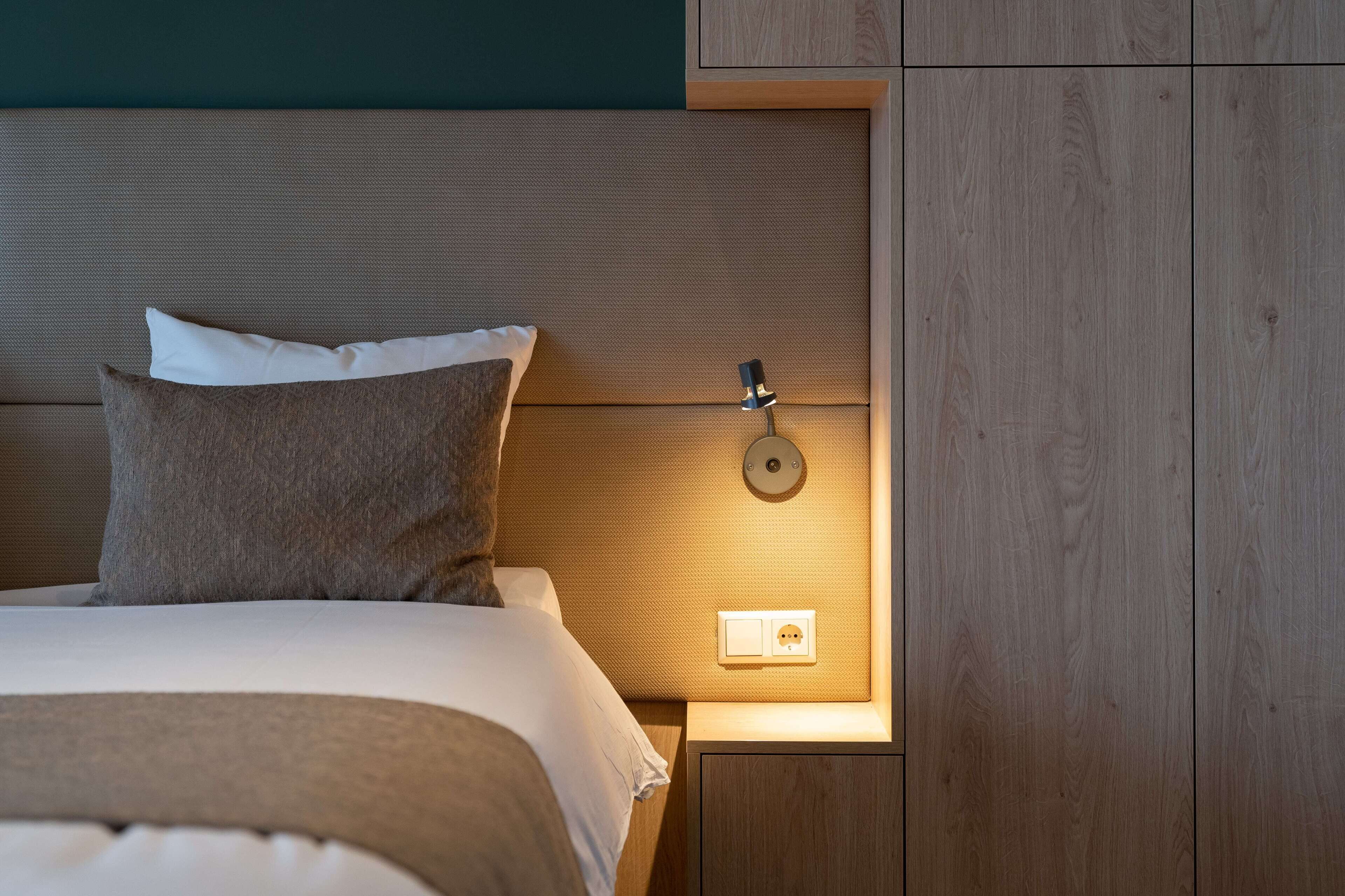 Un lit bien fait, avec une tête de lit, une lampe de chevet et une prise électrique sur une table de chevet en bois.