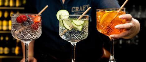 Tres cócteles con adornos, una persona sosteniendo uno, y una barra con botellas al fondo.