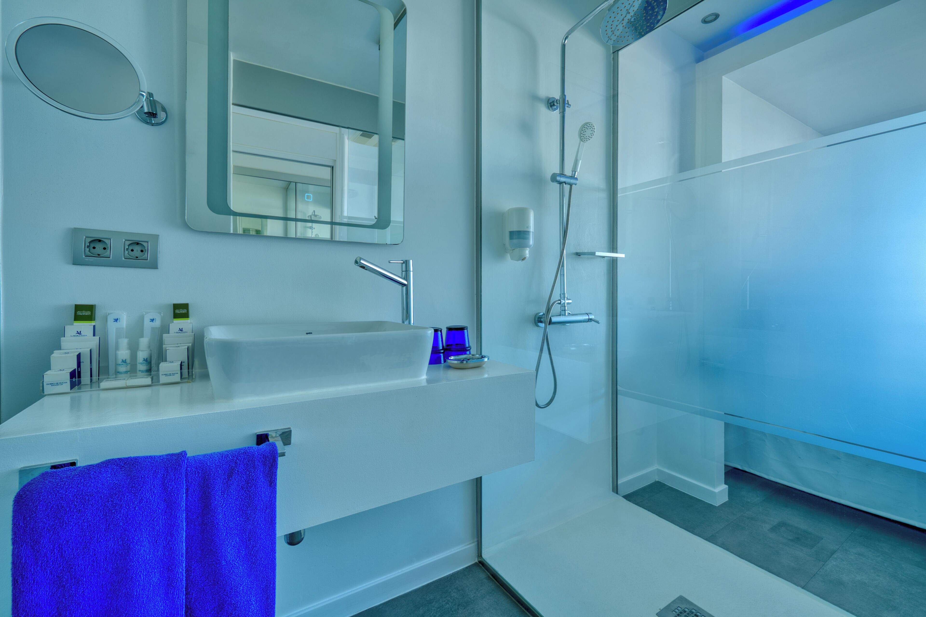 Une salle de bain moderne avec un grand miroir, un lavabo avec des articles de toilette, une douche et une serviette bleue.