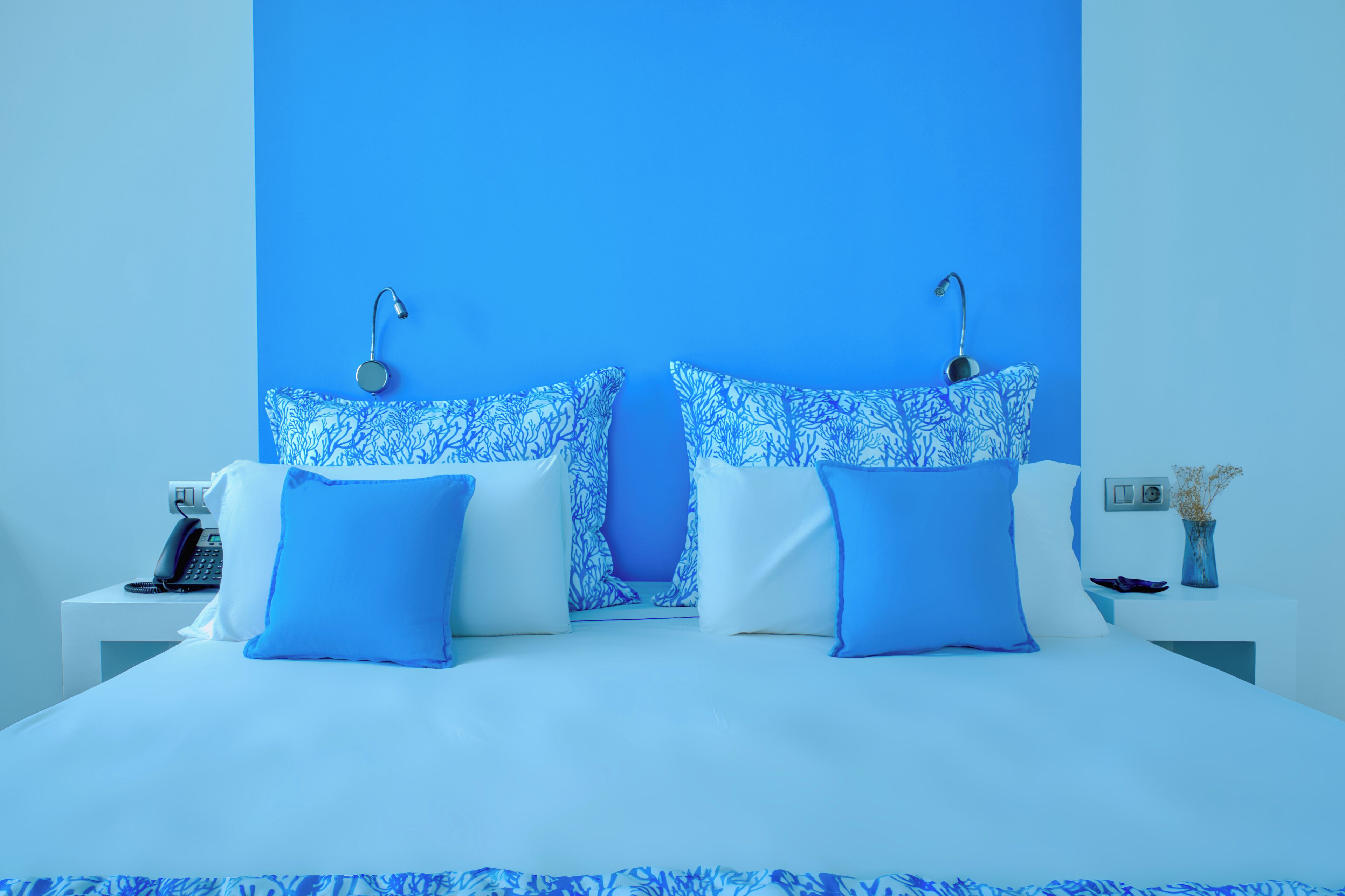 Una habitación de hotel moderna con una cama grande, dos lámparas de noche, una mesita de noche con un teléfono y un jarrón con flores secas.
