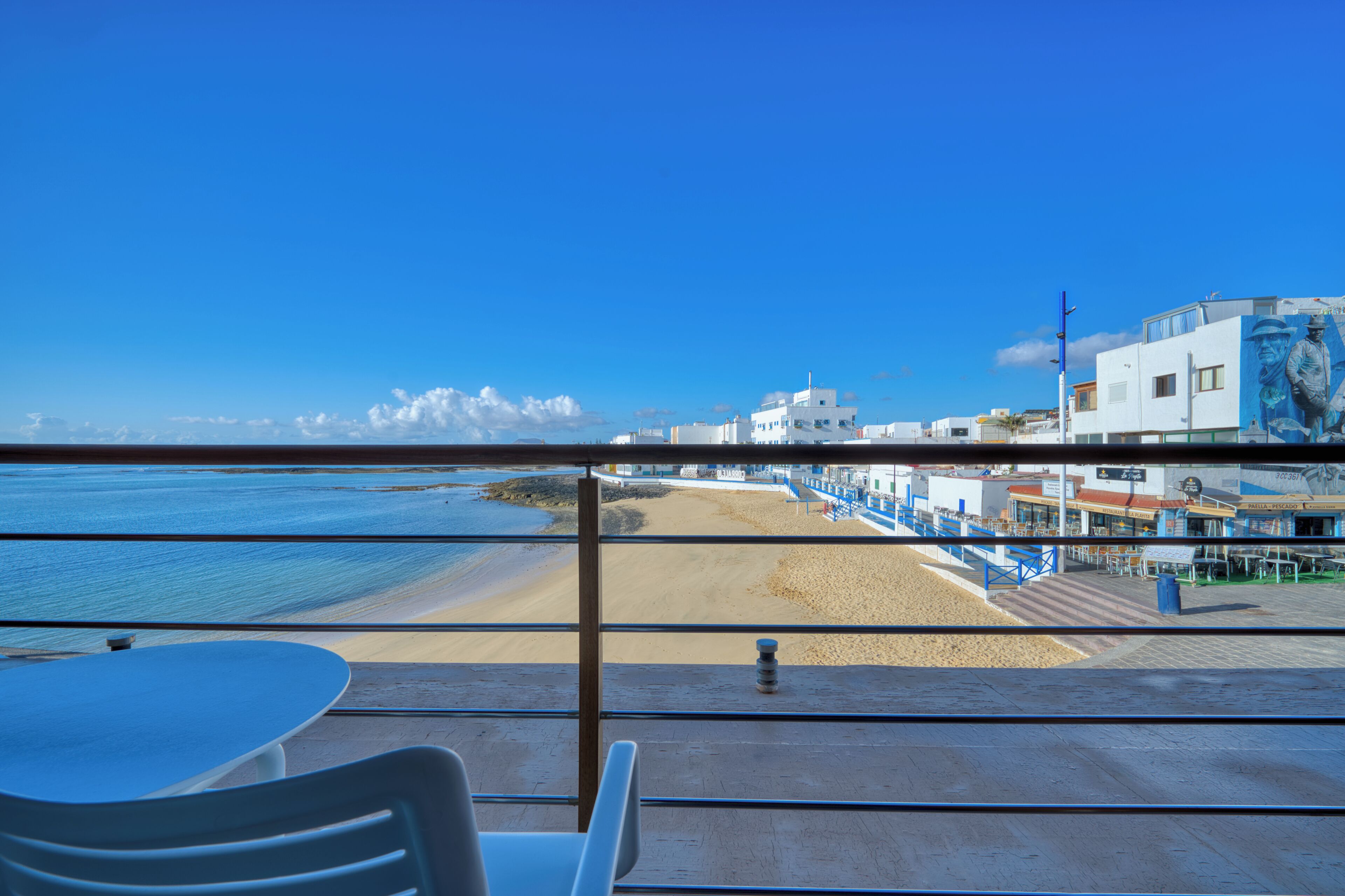 Blick vom Balkon auf einen Strand mit Tisch und Stühlen, ein sandiger Strand und ein Gebäude mit Wandgemälde.