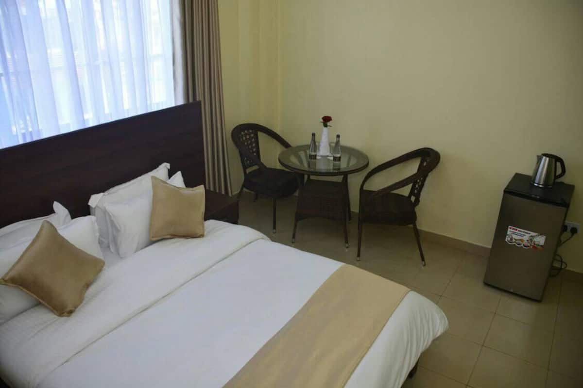 Quarto de hotel com cama, uma mesinha com cadeiras, minibar e janela com cortinas.