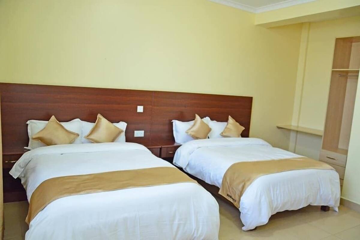 Quarto de hotel com duas camas, cabeceira de madeira, paredes bege e um guarda-roupa.