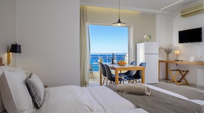 A hotel room with a bed, a desk with a chair, a dining table with chairs, a TV, and a view of the sea. - Bellevue Suites (Agios Nikolaos)