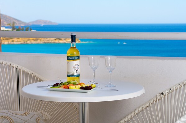 A table with a bottle of Enotria wine, a plate of fruit, and two empty wine glasses on a balcony with a view of the sea.