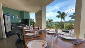 Un comedor con una larga mesa dispuesta para seis personas, una cocina al fondo y vista a un paisaje exuberante.