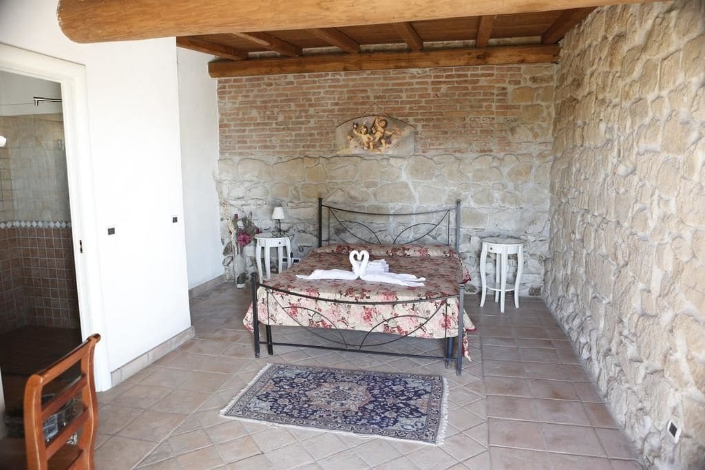 A bedroom with a stone wall, a bed with a floral bedspread, a small table with a lamp, and a chair.