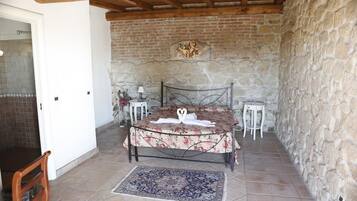 A bedroom with a stone wall, a bed with a floral bedspread, a small table with a lamp, and a chair.