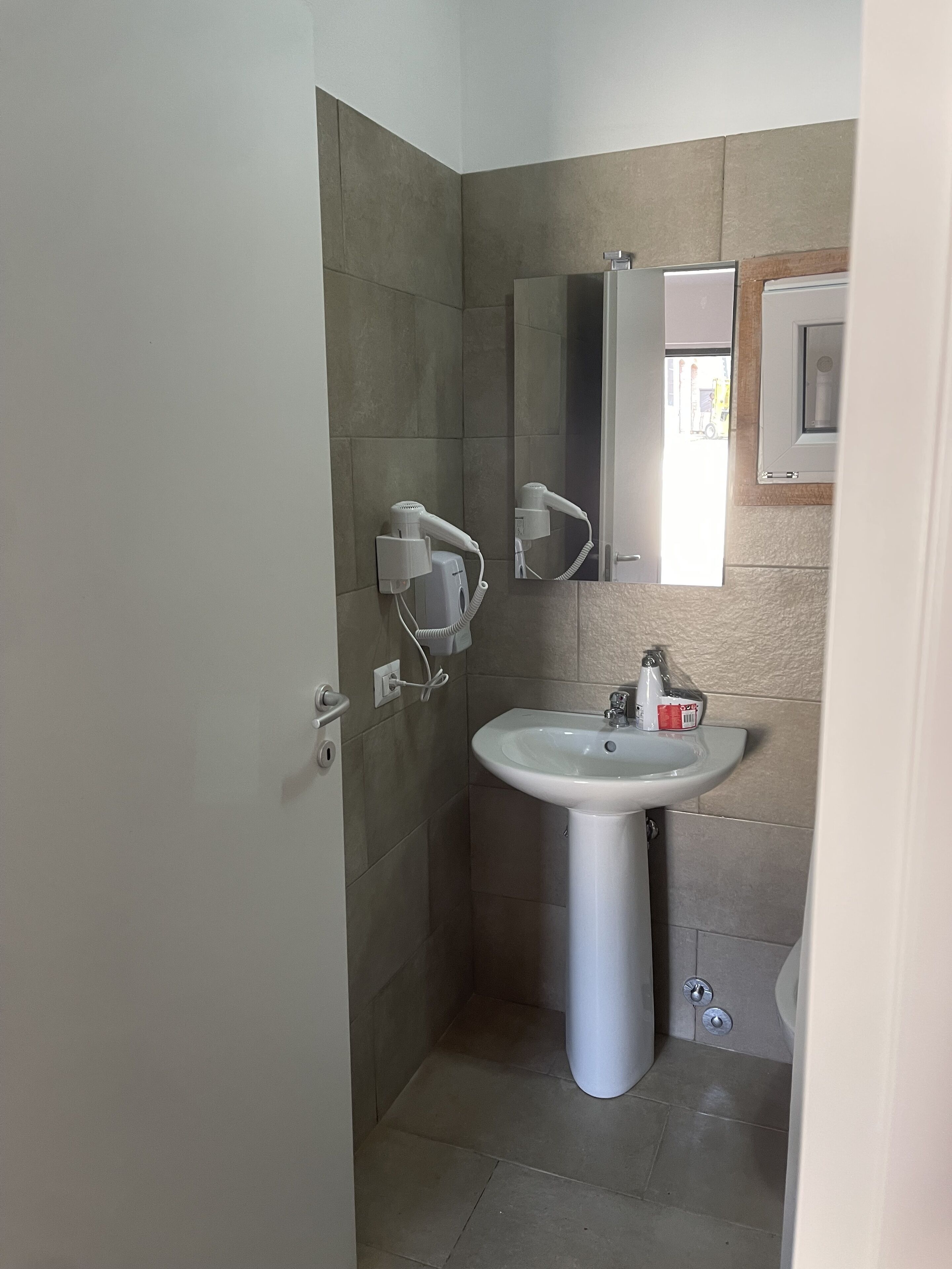 Uma casa de banho com lavatório pedestal branco, um espelho em cima e um secador de cabelo na parede.