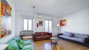 Amplia sala de estar con un sofá verde, un sofá de cuero marrón y un sofá gris estampado. Hay dos ventanas con marcos blancos, un radiador rojo y una lámpara de techo.