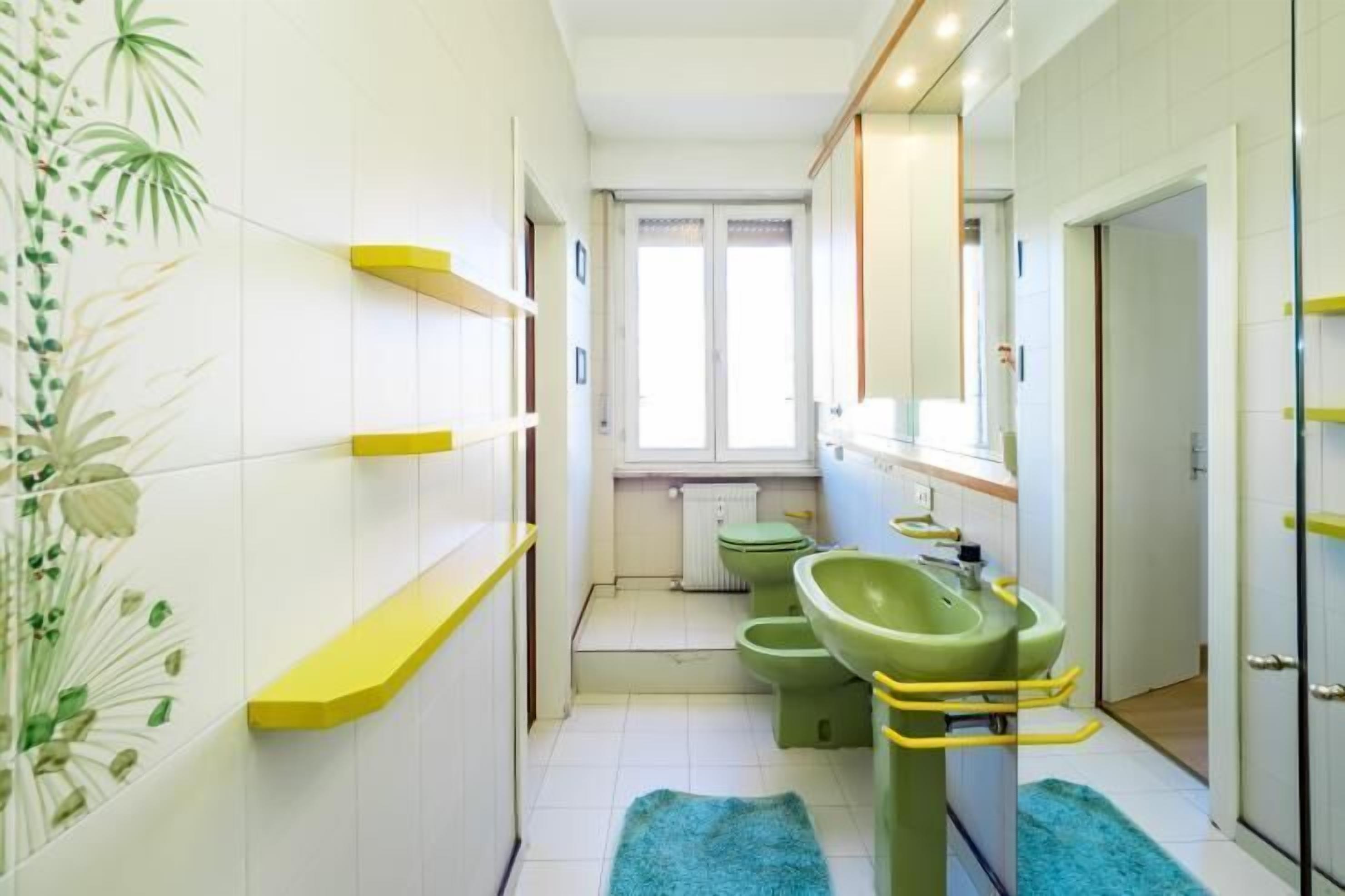 A narrow bathroom with white tiled walls, a green sink, and a toilet. There are yellow shelves and a blue rug on the floor.
