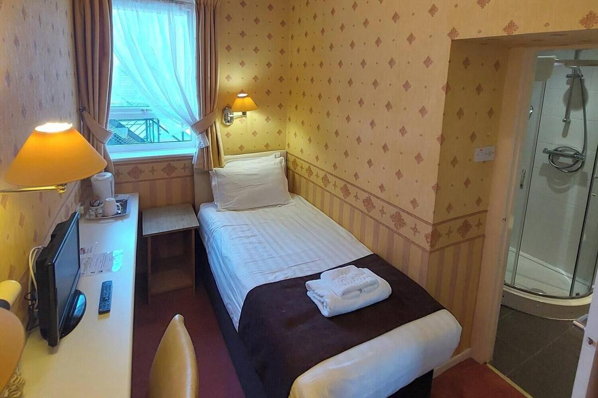 A hotel room with a bed, a desk with a lamp, a window with curtains, and a shower area.