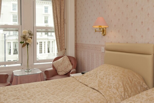 Habitación de hotel con cama, una silla, una mesita con un florero y una ventana con cortinas.