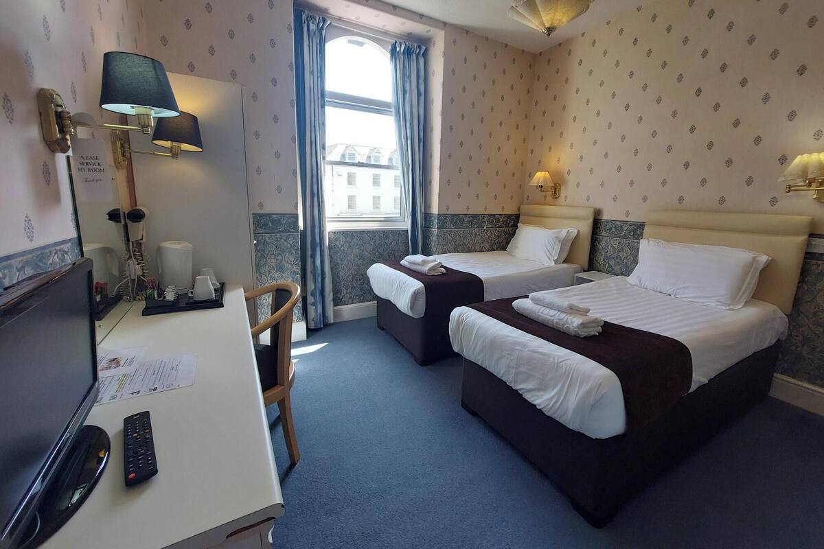 A hotel room with two beds, a desk with a lamp, a chair, and a window with curtains.