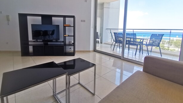 Una sala de estar moderna con televisor, mueble de entretenimiento negro y sofá. Hay un balcón con mesa y sillas, que ofrece vistas al mar.