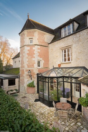 Un bâtiment en pierre doté d’une véranda en verre, d’une tour en briques et d’un patio en pierre avec une table et des chaises.