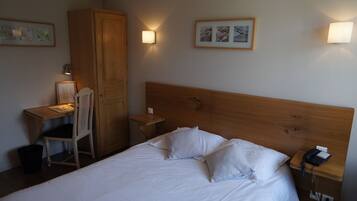 Quarto de hotel com cabeceira de madeira, cama com roupa de cama branca, escrivaninha com cadeira e duas luminárias fixadas na parede.