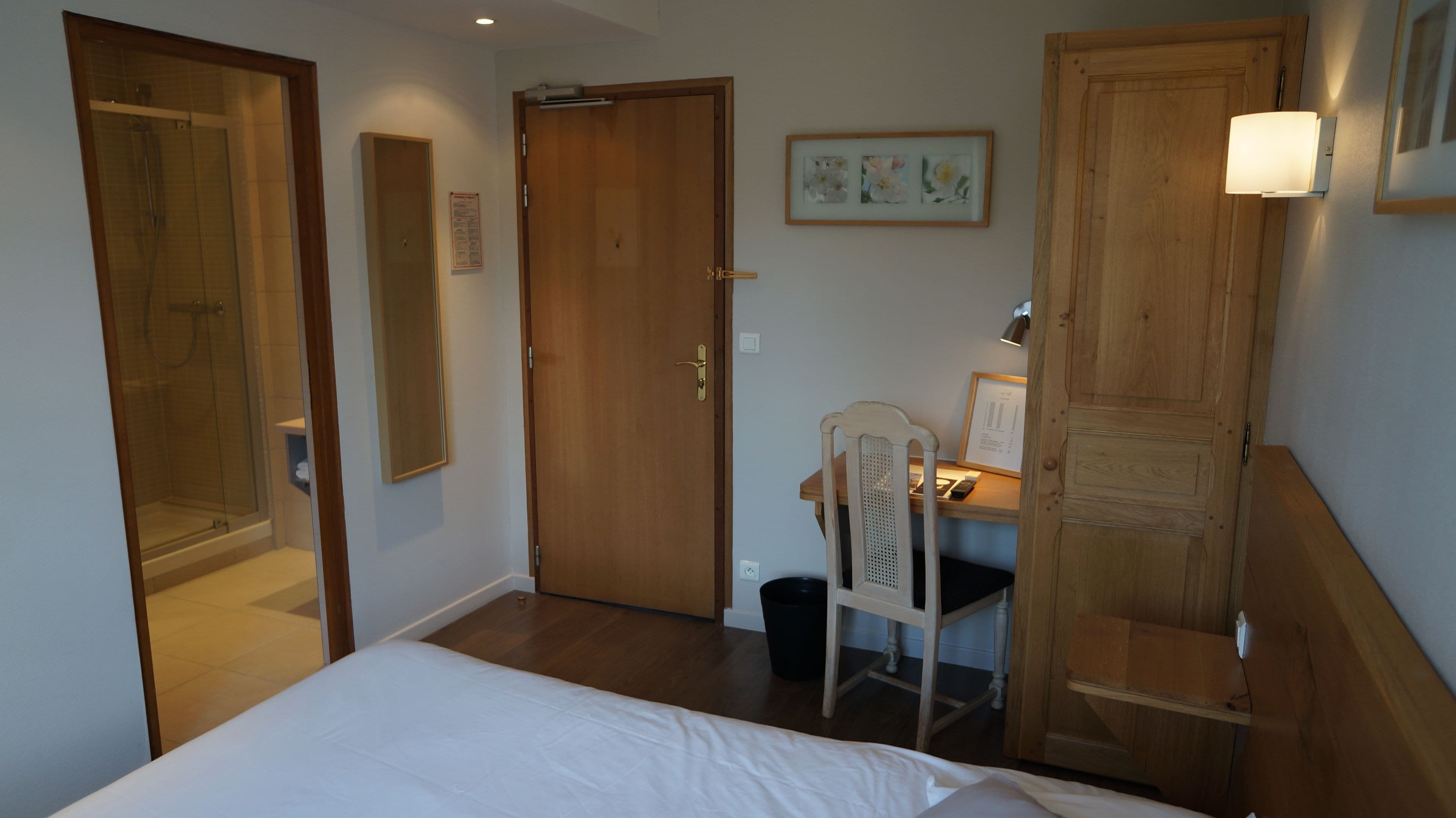 A hotel room with a bed, a desk with a chair, a wooden wardrobe, a framed picture on the wall, and a shower area.