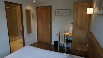 A hotel room with a bed, a desk with a chair, a wooden wardrobe, a framed picture on the wall, and a shower area.
