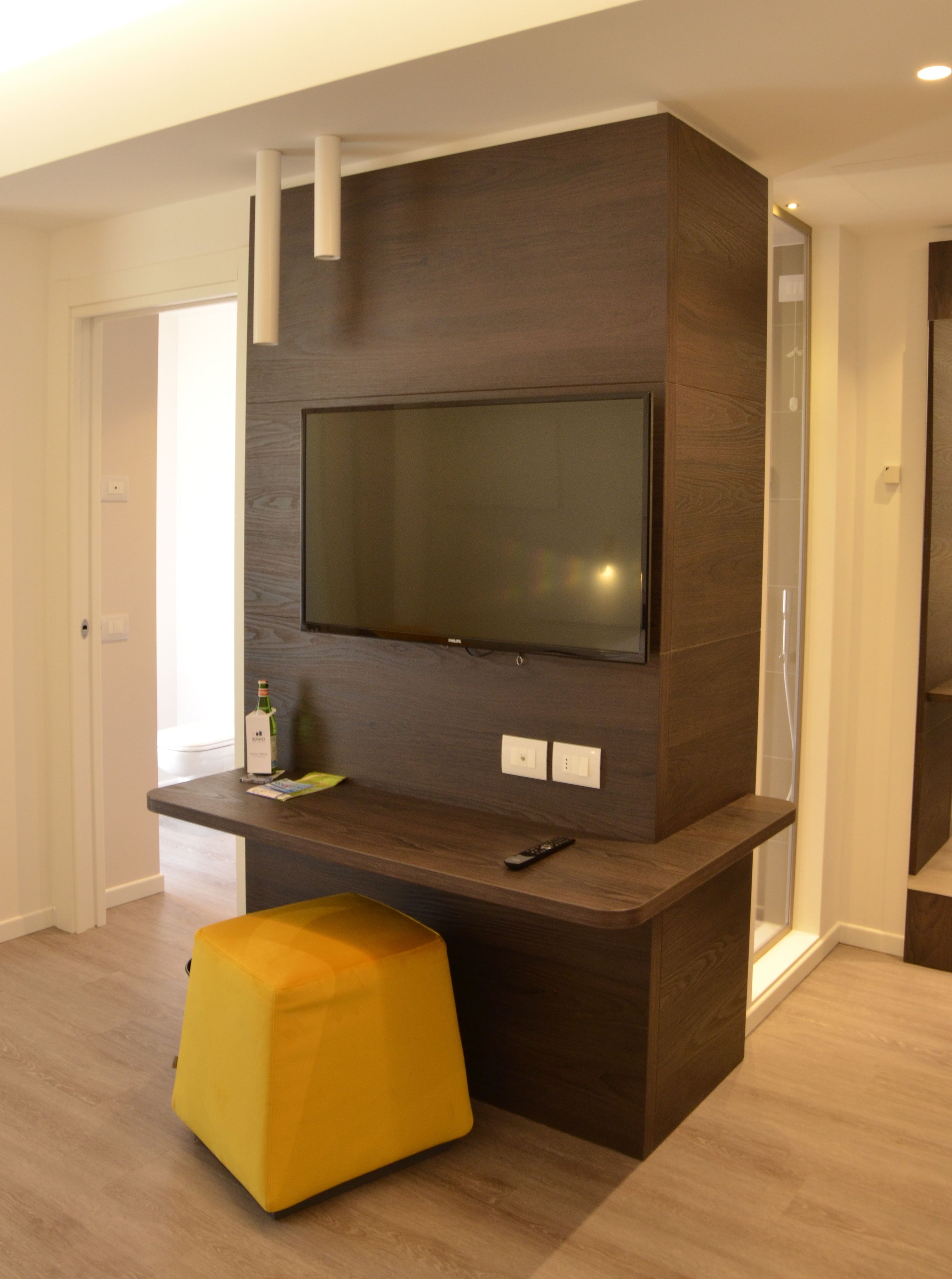 Una sala de estar moderna con un televisor de pantalla plana grande montado en una pared de madera oscura. Hay un otomano amarillo en primer plano y un control remoto sobre el mueble de televisión.