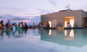 Un espace aménagé au bord de la piscine, avec un bar et des sièges, entouré d’un bâtiment moderne doté d’un toit plat et d’une antenne visible.