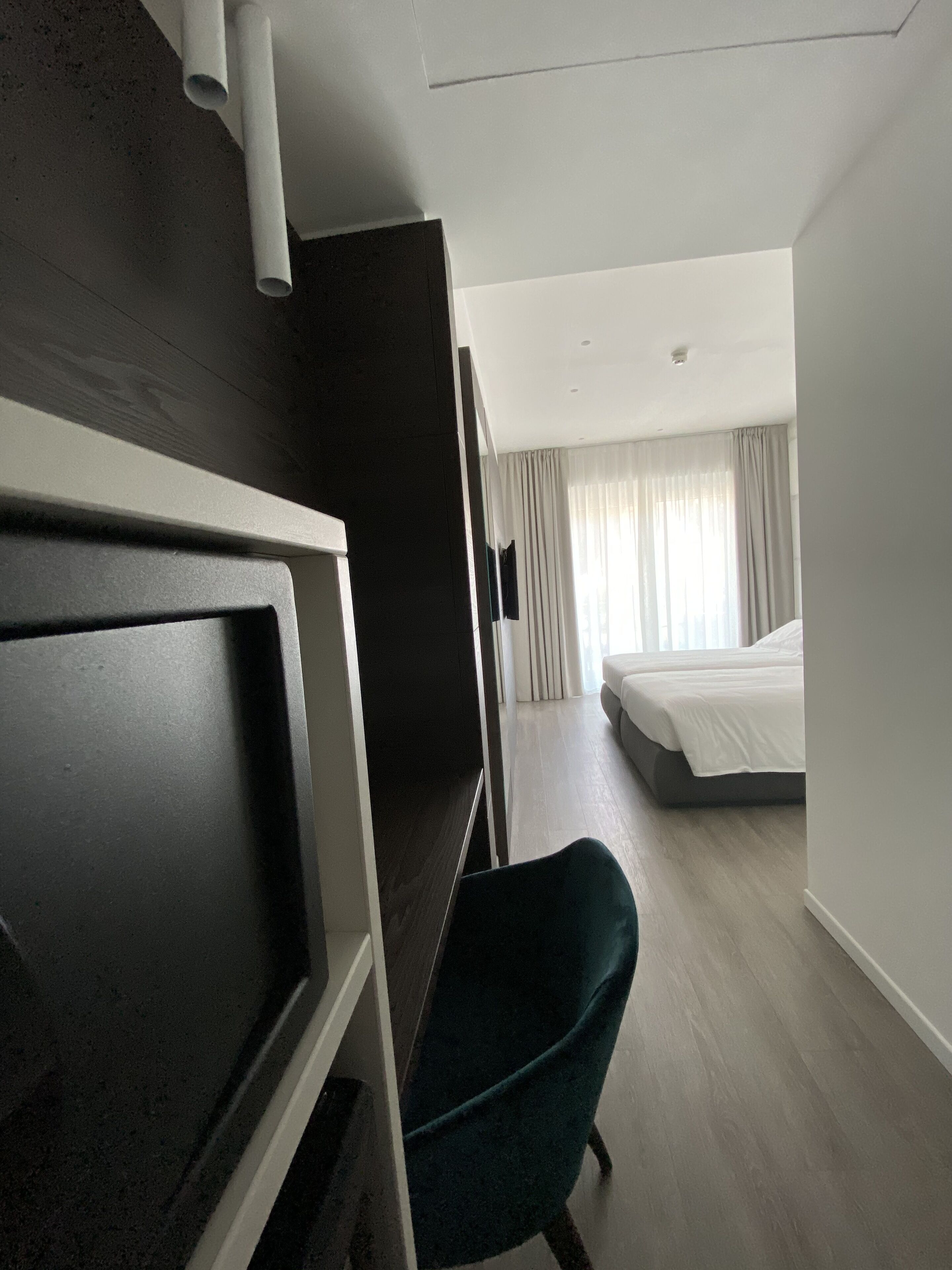 Habitación de hotel con un armario de madera oscura, una silla color turquesa, una cama con ropa de cama blanca y un ventanal con cortinas de gasa.