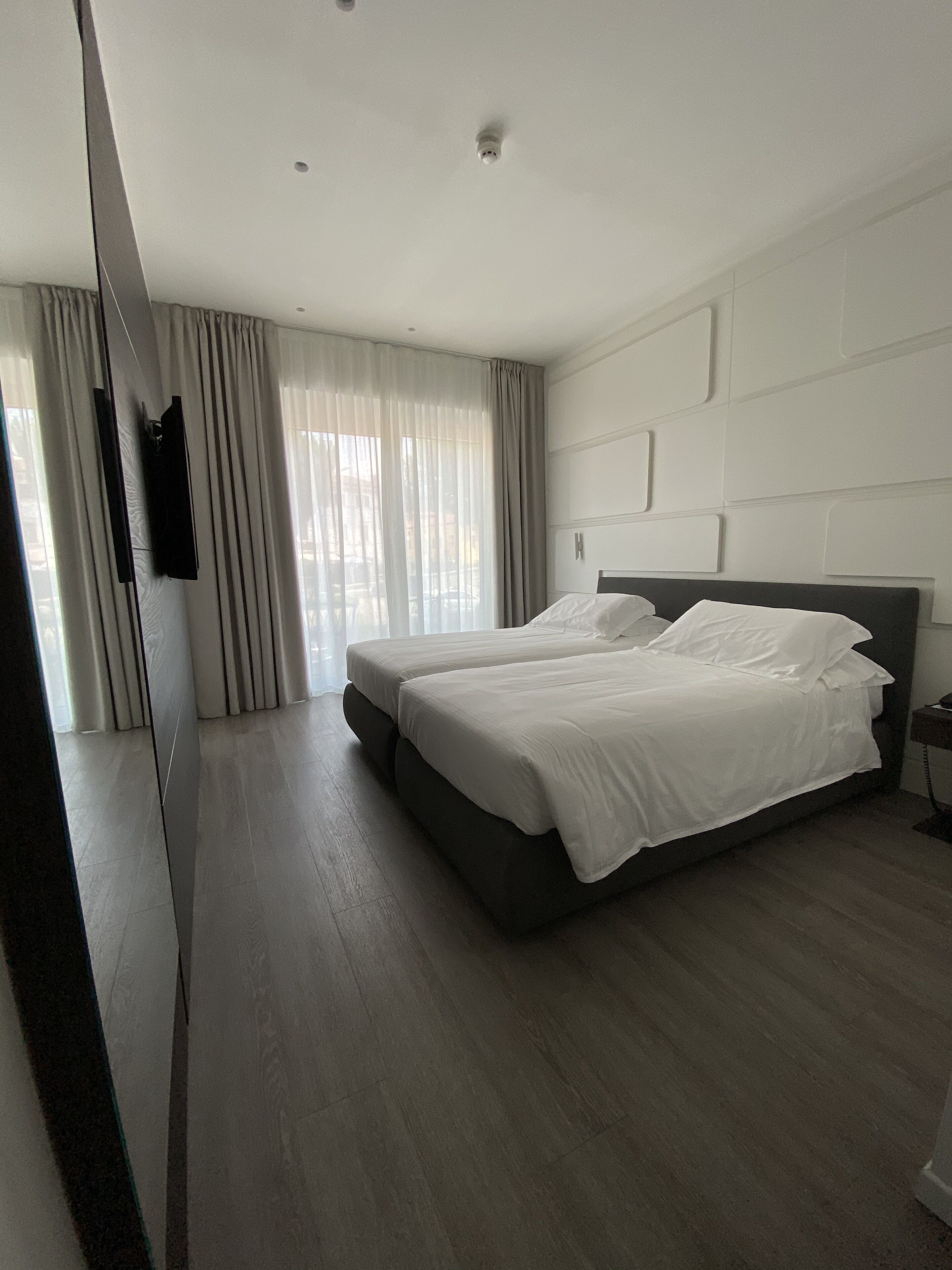 A modern hotel room with a large bed, a flat-screen TV mounted on the wall, and a large window with curtains.