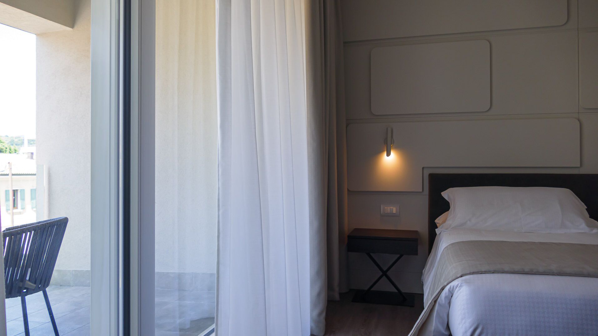 Une chambre d’hôtel avec un lit, une table de chevet, une lampe, une chaise et une fenêtre avec des rideaux.