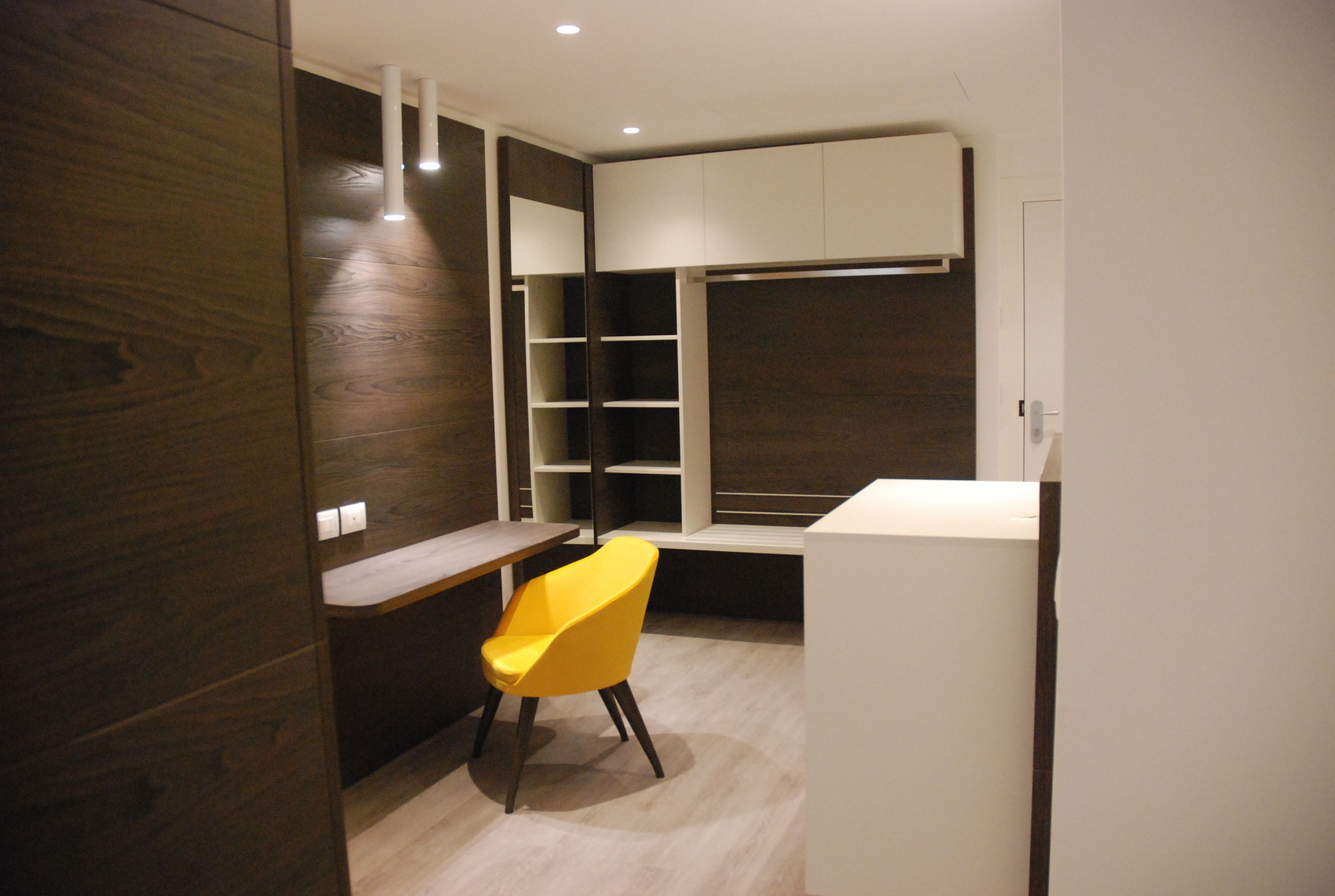 Une chambre moderne équipée d’un bureau, d’une chaise jaune, d’étagères intégrées et d’une armoire blanche.