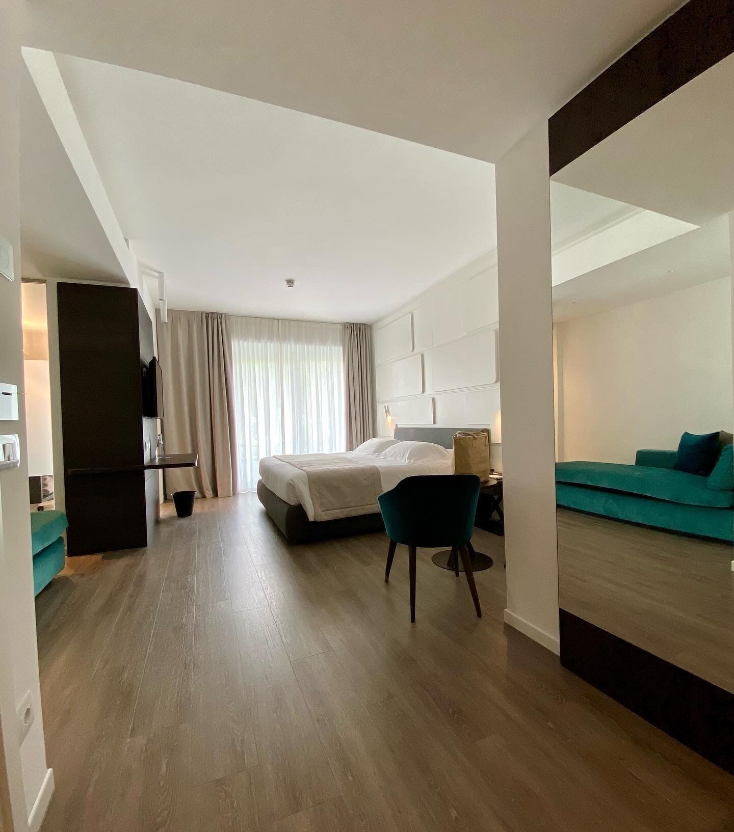 Ein modernes Hotelzimmer mit Bett, Schreibtisch, Sessel, Sofa und einem großen Spiegel.