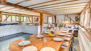 A spacious dining area with a long wooden table set for a meal, white cabinetry, and a high ceiling with exposed beams. - Butley Barn (Woodbridge)