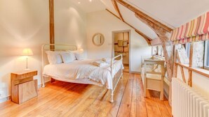 A bedroom with a wooden floor, a white bed, a bedside table, a radiator, and a window with striped curtains. - Butley Barn (Woodbridge)